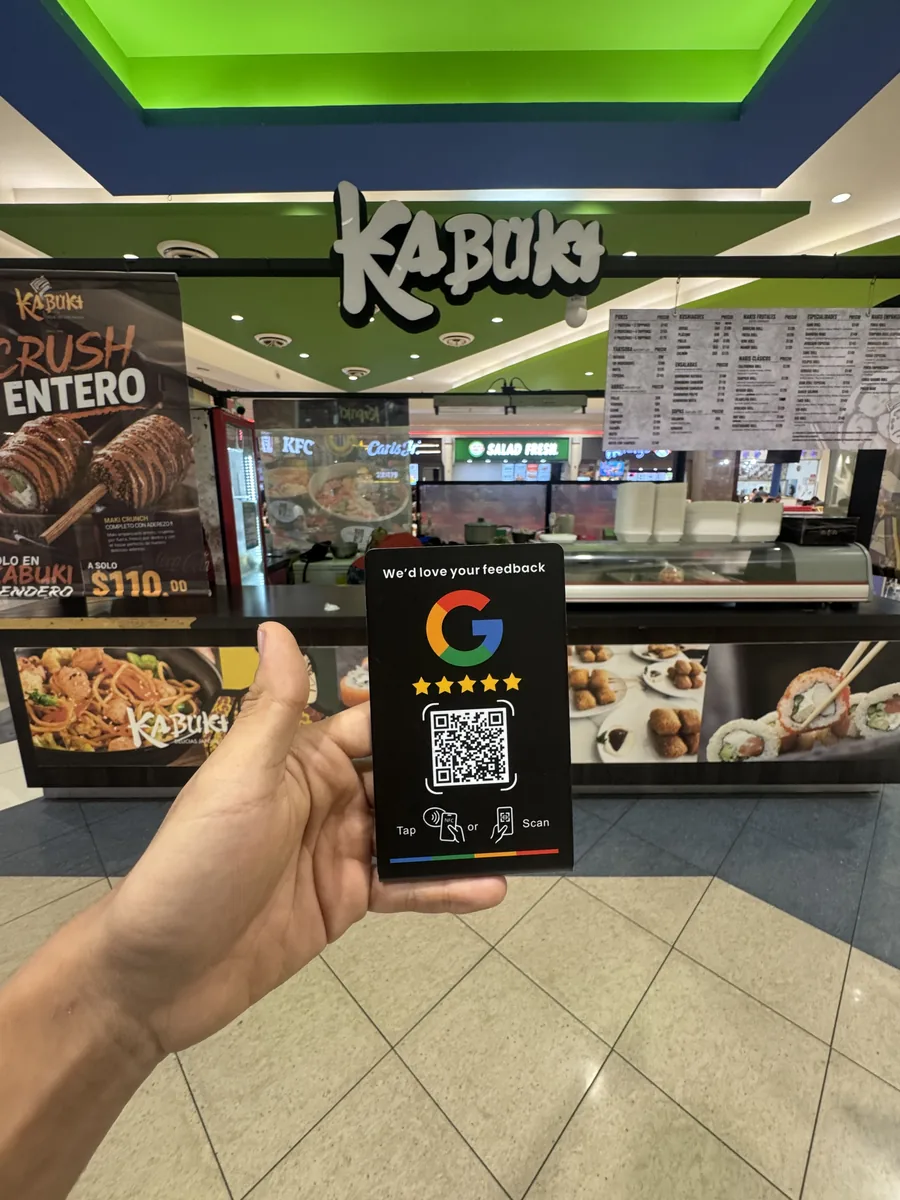 La placa de reseñas sostenida en la mano frente al mostrador de Kabuki en Plaza Sendero: al fondo se ven el rótulo del local, la carta y las vitrinas de sushi.