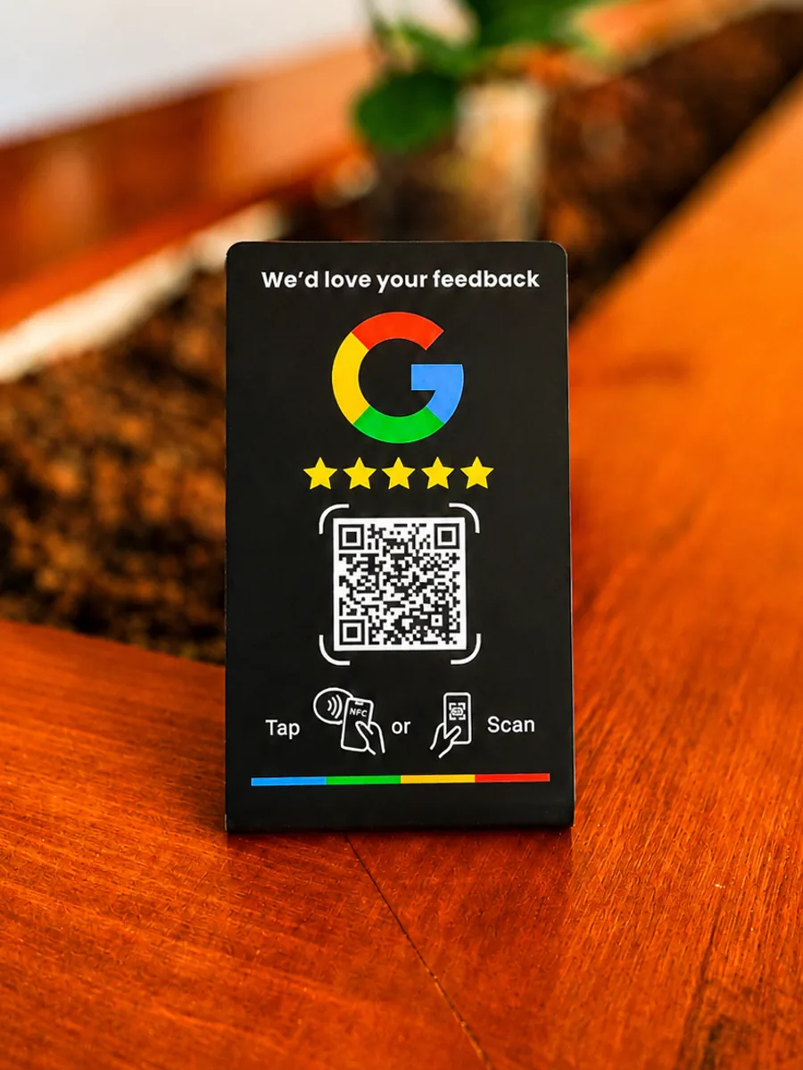 La placa de reseñas vista de frente sobre una mesa de madera: placa negra con el texto «We’d love your feedback», el logotipo de Google en sus cuatro colores, cinco estrellas doradas, un código QR y la indicación «Tap o Scan».